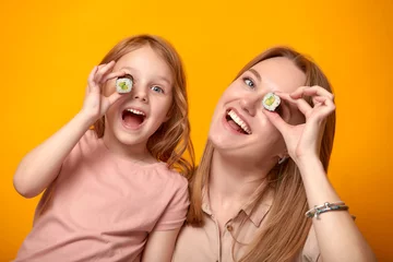  Funny mom with daughter holding sushi rolls in front of eyes on yellow background © Georgii