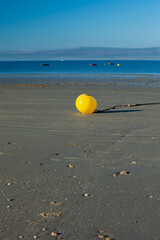 A yellow buoy on the sand at low tide. a yellow buoy contrasts sharply with the blue of the sky and the sea. Boats are moored behind.