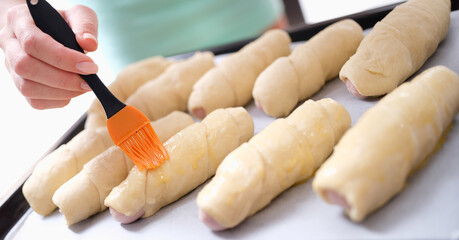 Female cook greases sheet of dough with melted butter and prepares sausage wrapped in dough