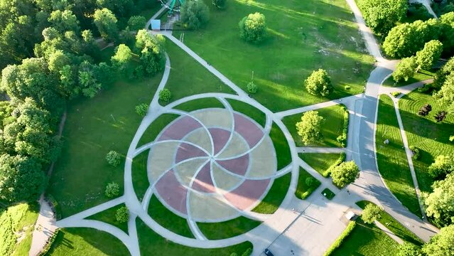 In a city park covered with trees and grass, with paths for walking, in the middle of the park there is a circle paved with colored pavers.