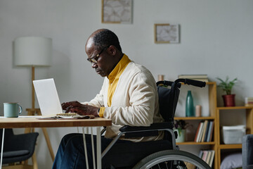 African man in eyeglasses sitting on wheelchair at table and typing on laptop at home