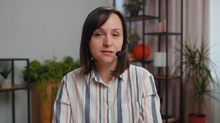 Young business woman sitting at desk at home office wearing headset, freelance worker, call center or support service operator helpline, having talk with client or colleague, communication support