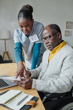 African Female Volunteer Pointing To Receipt And Explaining Man With Disability How To Pay Online On Laptop