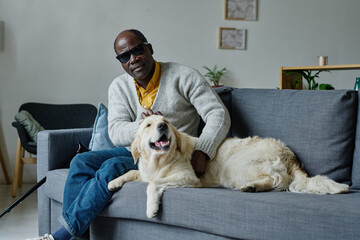 Portrait of African blind man in dark glasses resting on sofa in living room together with his...