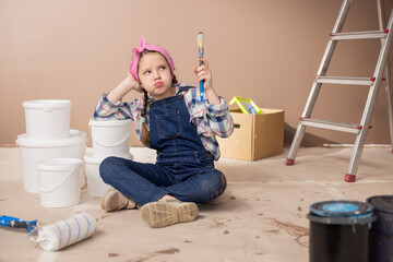 Naklejka premium A sad tired girl props elbow on the paint cans sighs lets the air out of mouth with an expression of lack of strength, holds in hand a paintbrush soiled with the brown color of the walls.