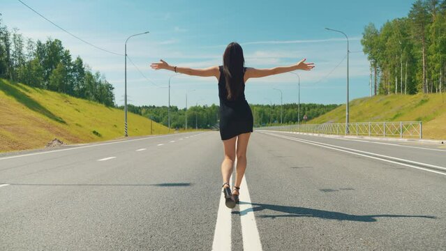 rear view of a girl in a tight black dress walking in the middle of a highway on a sunny day. The girl throws her hands up, welcoming the sun and freedom
