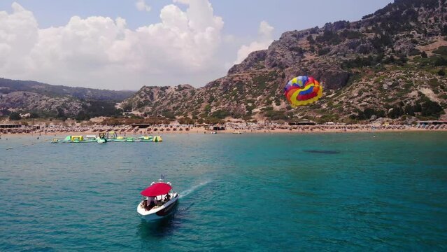 Parasailing - Speedboat Towing Tourists Attached On Colorful Parachute In Tsambika Beach, Rhodos, Greece. - aerial