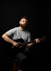 Bearded brutal guitarist plays an acoustic guitar in a black room and looking in the camera. The concept of music recording, rehearsal or live performance.