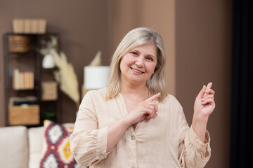 A cheerful elderly middle-aged woman stands in the home's warm living room. Retired woman smiles points upward above her,,gestures, advertising space caption.