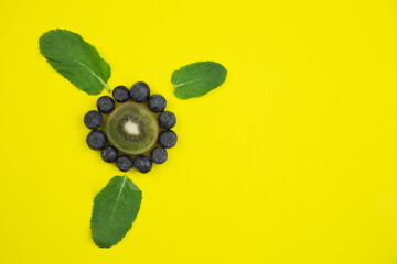 A creative flower consisting of a cut kiwi with a heart and petals in the form of blueberries, decorated with mint leaves lies on the side on a bright yellow background with a place for text. High