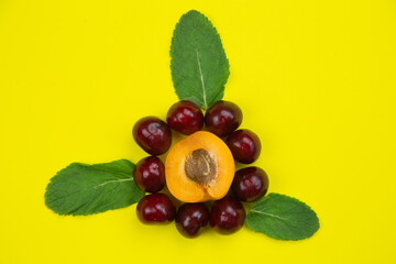 A creative flower consisting of half an apricot with a heart stone and petals in the form of cherries, decorated with mint leaves lies in the middle on a bright yellow background. High quality photo