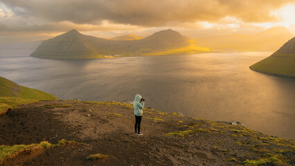 Lonely tourist in blue jacket looking over majestic fjords on Faroe Islands. Landscape photography. Great sunset view from populat tourist attraction - Klakkur peak