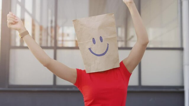 A crazy anonymous woman is gesturing with a cardboard box on his head with smiley face. Funny person is doing winner gesture and celebrating. Concept of victory or happiness and freedom.