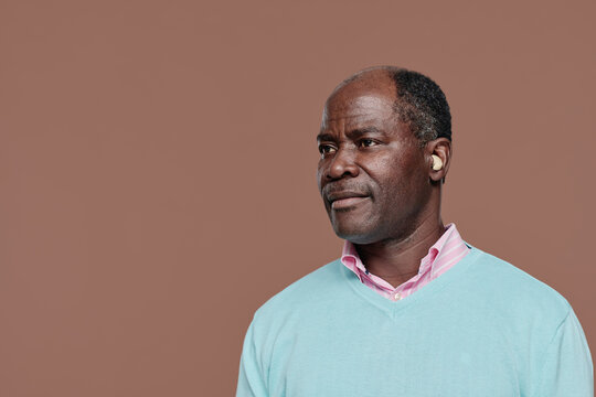 Portrait of senior deaf African man with hearing aid standing against brown background