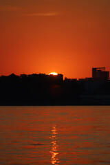 Fototapeta premium A bright setting sun sets behind the black silhouettes of buildings above the river against an orange red sky. Summer sunset background. Beautiful landscape, close up