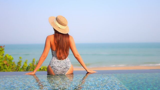 Back View of Woman With Fit Body, Floppy Hat in Swimsuit Sitting on Pool Edge and Looking at Tropical Beach and Sea Horizon