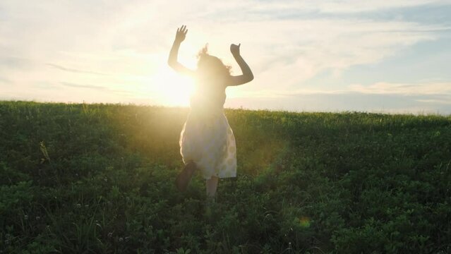 Girl child in a dress runs across the grass to meet the setting sun and waves her arms like a bird flapping her wings. Slow motion. Concept of childhood dreams, freedom, unity with nature. 