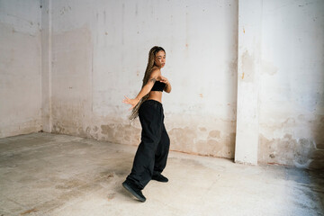 multiracial girl with long blonde braids dancing in a black tracksuit in an abandoned warehouse while smiling and enjoying her passion and exercise.