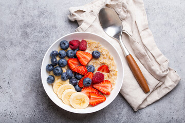 Oatmeal porridge with fruit and berries in bowl with spoon on gray stone background table top view, homemade healthy breakfast cereal with strawberry, banana, blueberry, raspberry