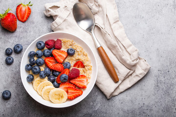 Oatmeal porridge with fruit and berries in bowl with spoon on gray stone background table top view, homemade healthy breakfast cereal with strawberry, banana, blueberry, raspberry. Space for text