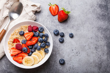 Oatmeal porridge with fruit and berries in bowl with spoon on gray stone background table top view, homemade healthy breakfast cereal with strawberry, banana, blueberry, raspberry. Space for text