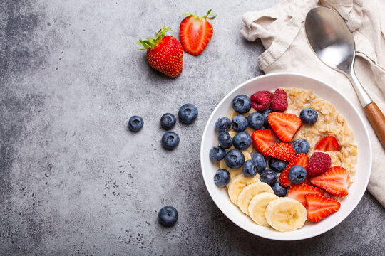 Oatmeal porridge with fruit and berries in bowl with spoon on gray stone background table top view, homemade healthy breakfast cereal with strawberry, banana, blueberry, raspberry. Space for text