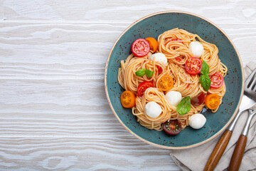 Delicious spaghetti with mozzarella, colourful cherry tomatoes, fresh basil on ceramic plate rustic white wooden background top view. Italian healthy tasty food pasta for dinner or lunch. Copy space