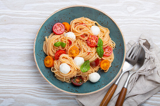 Delicious spaghetti with mozzarella, colourful cherry tomatoes, fresh basil on ceramic plate rustic white wooden background top view. Italian healthy tasty food pasta for dinner or lunch