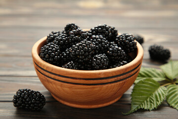 Ripe blackberries with leaves in a wooden bowl on brown wooden background.