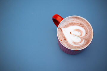 Mug of hot chocolate with latte art in the shape of a heart on a blue surface. Top view