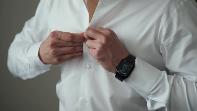 Groom buttoning his white shirt before wedding ceremony alone standing by the window with black smart watch on his wrist.
