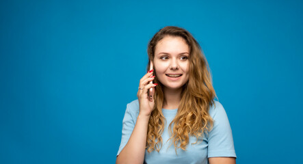 Fototapeta premium Portrait of a surprised girl with a curly hair dressed in blue t-shirt talking on mobile phone isolated over blue background.