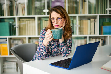 Businesswoman working on laptop in the library and holding a cup of coffee 