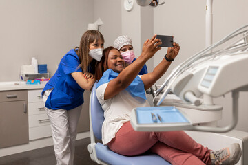A black woman patient, takes a happy selfie photo with the smartphone side by side the dentist and dental hygienist at the dental clinic