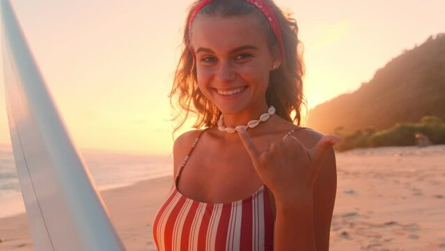 Young beautiful Caucasian woman teenager cute smiling holding surfboard and showing fingers shaka gesture getting ready to swim on sea waves relaxing on summer weekend stands on coast of ocean