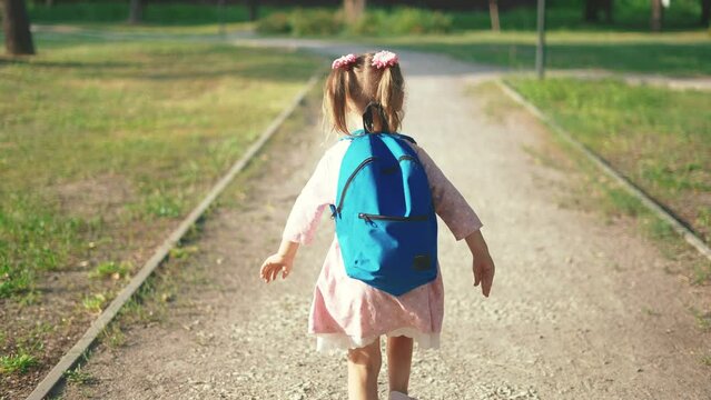 back to school. a little girl with a backpack runs through the green park to school along the path. education kid dream concept. a small child sun with a backpack back view runs the park to school