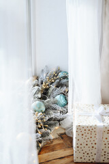 Snow-covered Christmas Tree with Blue Balls on Window. White Curtains in New Year's Interior. Gift