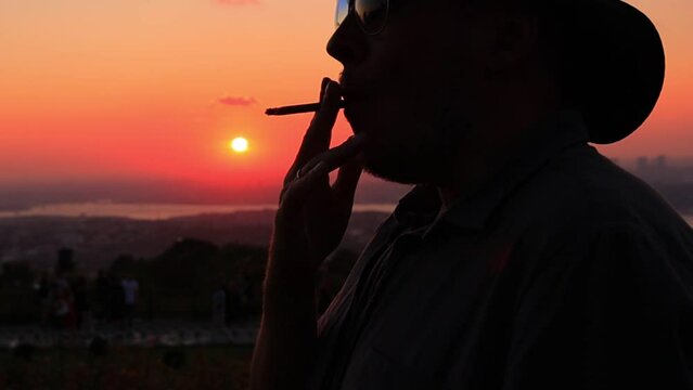 Silhouette man in a hat smoking a small cigar at an orange sunset in the evening