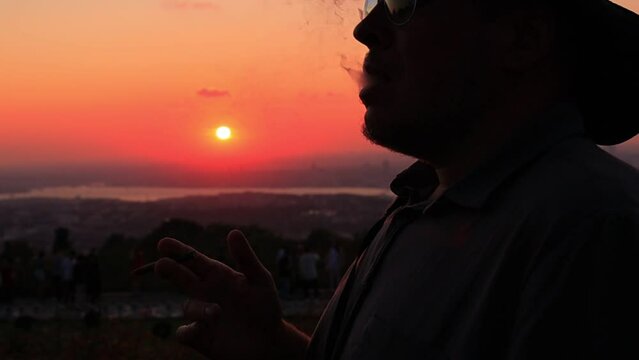 Silhouette man in a hat smoking a small cigar at an orange sunset in the evening