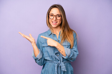 pretty young adult woman smiling cheerfully and pointing to copy space on palm on the side, showing...