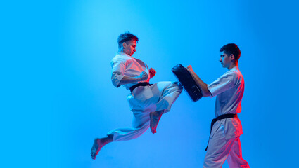 Studio shot of sports training of two karatedo fighters in doboks isolated on blue background in neon. Concept of combat sport, challenges, skills