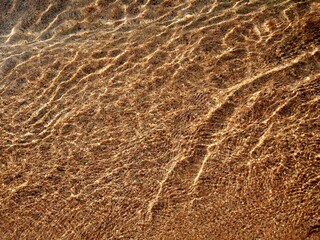 Sea water in shallow water, which can be seen in shallow water - large pebbles, east coast of Spain. The water sparkles and shimmers in the sun. Clear sunny day at the sea. Photo in shades of measles 