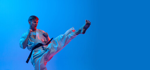 Fototapeta na wymiar One young man, karate fighter in white kimono in action, motion isolated on blue background in neon. Sport, action, combat sports, ad concept