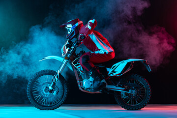 Portrait of young man, biker in full equipment riding motorbike isolated over dark studio...
