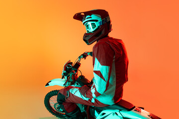 Portrait of young man, biker in full equipments posing on motorbike isolated over orange studio...