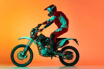 Fototapeta na wymiar Portrait of young man, biker in full equipments riding motorbike isolated over orange studio background in neon light. Doing tricks
