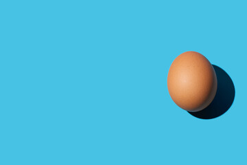 A brown chicken egg lies on the left side of the surface on a blue background. Hard light, deep shadow, horizontal. Close-up, copy space, top view.