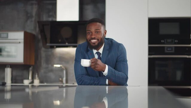 African-American man in suit drink coffee and smile standing in modern kitchen. Realtime
