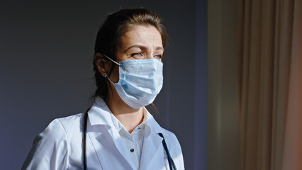 Charismatic mature woman doctor take her protective mask and wearing on her face and looking through the hospital panoramic window