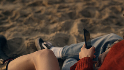 Couple legs sitting on sandy beach. Trendy girl holding smartphone in hand